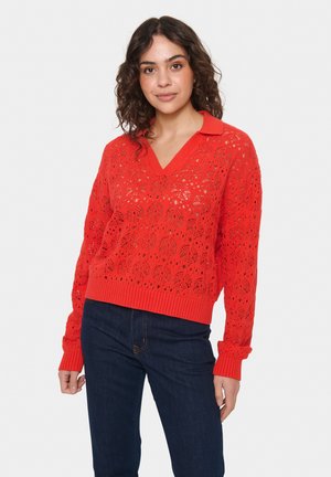 Maglione rosso lavorato a maglia con scollo a V e colletto, caratterizzato da un motivo in pizzo floreale e polsini a costine, abbinato a jeans blu scuro.