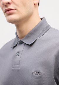 Siva polo majica od glatkog materijala, s ovratnikom, dvostrukim dugmadima i izvezenim "BOSS" logotipom na prsima.