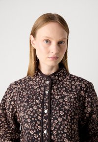 Woman with straight blonde hair wearing large hoop earrings and a dark floral buttoned jacket against a plain light background.