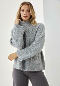 Suéter gris de punto con cuello alto, mangas anchas y patrón texturizado, combinado con pantalones vaqueros grises. Material suave y esponjoso con un ajuste relajado.