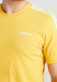 Camiseta de algodón amarilla con cuello redondo, que presenta el logo blanco de Adidas y tres rayas blancas a lo largo de los hombros. Textura suave.
