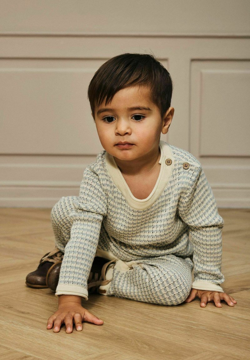 Un tout-petit portant une tenue légère bleu clair et crème à motifs, assis sur un sol en bois, regardant pensivement vers le bas.