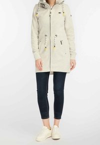 Un hoodie long gris clair avec une fermeture éclair, une capuche à cordon, des poches avant et du matériel contrasté noir et jaune. Associé avec un jean foncé et des baskets.