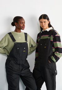 Zwarte denim salopettes met verstelbare banden en voorkeurszakken; het ene model in een lichtgroen sweatshirt, het andere in een gestreepte zwart-groene lange mouw.