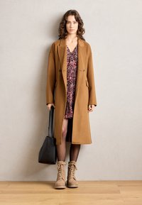 Abrigo de lana marrón con mangas largas, dos botones y bolsillos sobre un vestido rosa con flores. Bolso negro y botines beige con cordones.