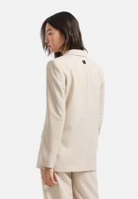 Blazer beige sartoriale con design strutturato, bottone singolo e un logo discreto sul retro. Tessuto liscio, forma aderente.