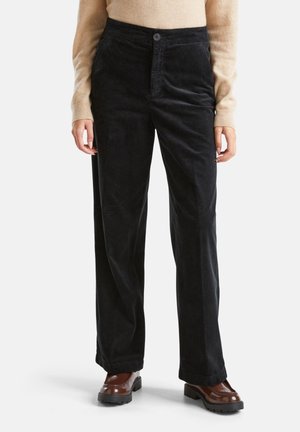 Femme portant un pantalon large en velours côtelé noir, un pull beige à manches longues et des mocassins en cuir marron sur un fond blanc.