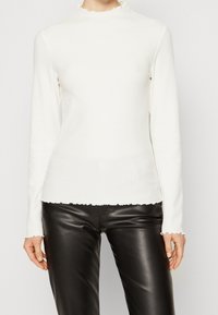 Maglietta bianca a maniche lunghe con texture a coste e colletto alto arricciato; abbinata a pantaloni in pelle nera.