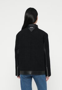 Μαύρο μπουφάν με υφασμάτινη επιφάνεια και δερμάτινα αξεσουάρ, με ψηλό γιακά και λεπτομέρειες με φερμουάρ στα πλάγια. Φορεμένο πάνω από μπλε τζιν.