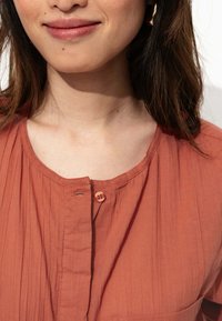 Chemise en tissu léger de couleur rouille, avec un col rond, des rayures verticales et une fermeture à boutons comportant deux boutons au niveau du col.