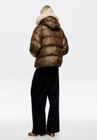 Hnedá púchrová bunda s prešívaným dizajnom a kapucňou s kožúškom, kombinovaná s čiernymi širokými nohavicami a vzorovanými topánkami.