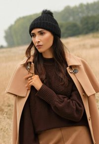 Căciulă neagră din tricot cu un pom-pom din blană sintetică, pulover texturat maroniu și palton bej cu nasturi mari și un design croit.