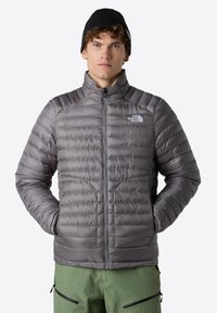 Šedá péřová bunda s horizontálním prošíváním a vysokým límcem, zapínání na zip, s logem na levé straně hrudi. Vytvořeno v kombinaci se zelenými kalhotami.