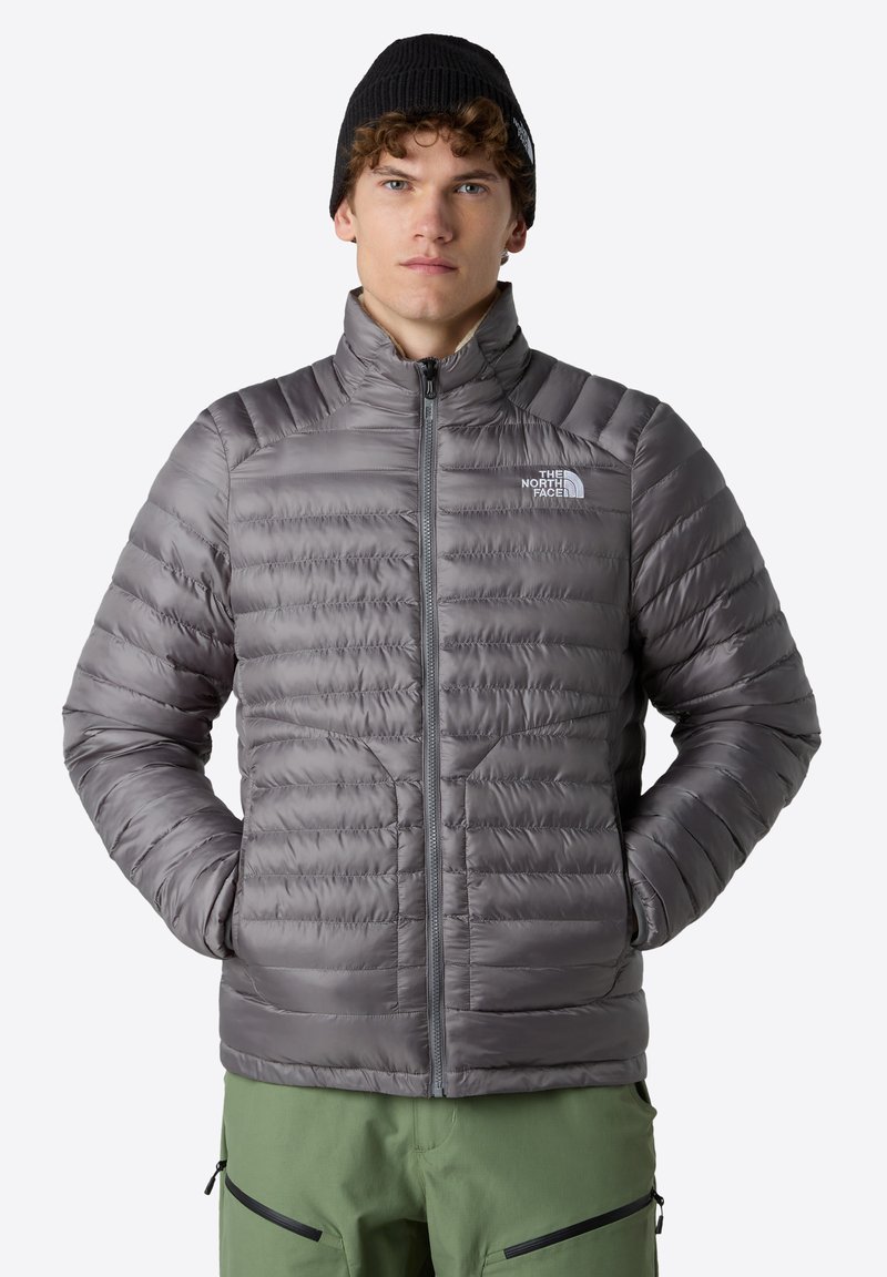 Šedá péřová bunda s horizontálním prošíváním a vysokým límcem, zapínání na zip, s logem na levé straně hrudi. Vytvořeno v kombinaci se zelenými kalhotami.