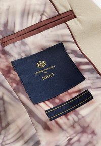 Etiketten har en mörkblå tygplatta med guldtexten "Designad och tillverkad av NEXT." Det omgivande tyget visar ett mjukt, blommönstrat mönster.