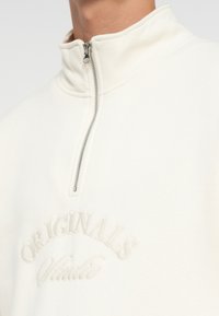 Pullover in pile bianco con zip a un quarto e il testo ricamato "ORIGINALS" sul petto. Texture liscia e dettagli di cucitura sottili.