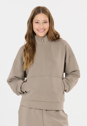 Junge Frau mit langen hellbraunen Haaren, die einen beigen Reißverschluss-Sweatshirt trägt und mit den Händen in den vorderen Taschen lächelt in die Kamera.