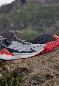 Szare i czarne buty do biegania z czerwonymi akcentami, z teksturowaną podeszwą, oddychającą cholewką z trzema białymi paskami i metką GORE-TEX.