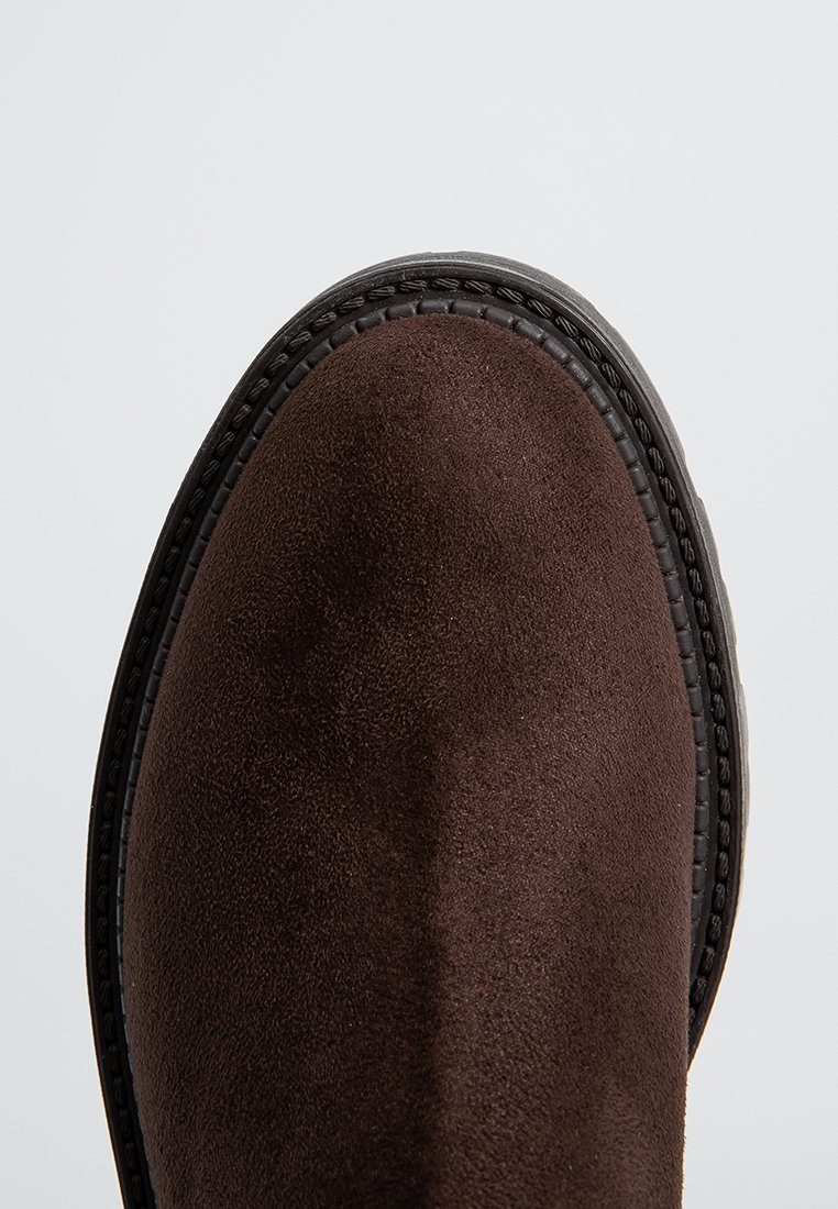 Stivale alla caviglia in suede marrone con punta arrotondata, superficie texturizzata e cuciture decorative lungo la suola in gomma scura. Profilo piatto.