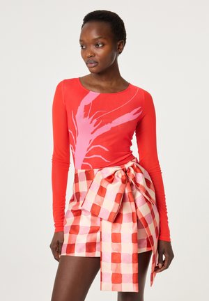 Vrouw draagt een rood longsleeve shirt met een roze kreeftenprint en een roze-rode geruite wikkelrok met een grote strik.