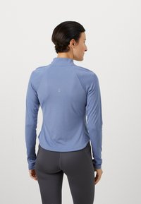 Lyseblå langermet treningsgenser med høy krage, med en teksturert bakside og sømløst design. Kombinert med mørke, tettsittende leggings.