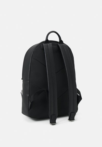 Musta reppu pyöreällä ylhäällä, pehmustetuilla mustilla hihnoilla ja hienovaraisella diagonaalisella ompeleella rakenteen vuoksi. Varustettu ylhäällä olevalla kahvalla ja sivutaskulla.