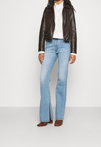 Frau in einer dunkelbraunen Lederjacke, weißer Rüschenbluse, hellblauen Jeans mit Schlitz vorne und braunen High-Heel-Stiefeln, stehend vor einem einfarbigen Hintergrund.