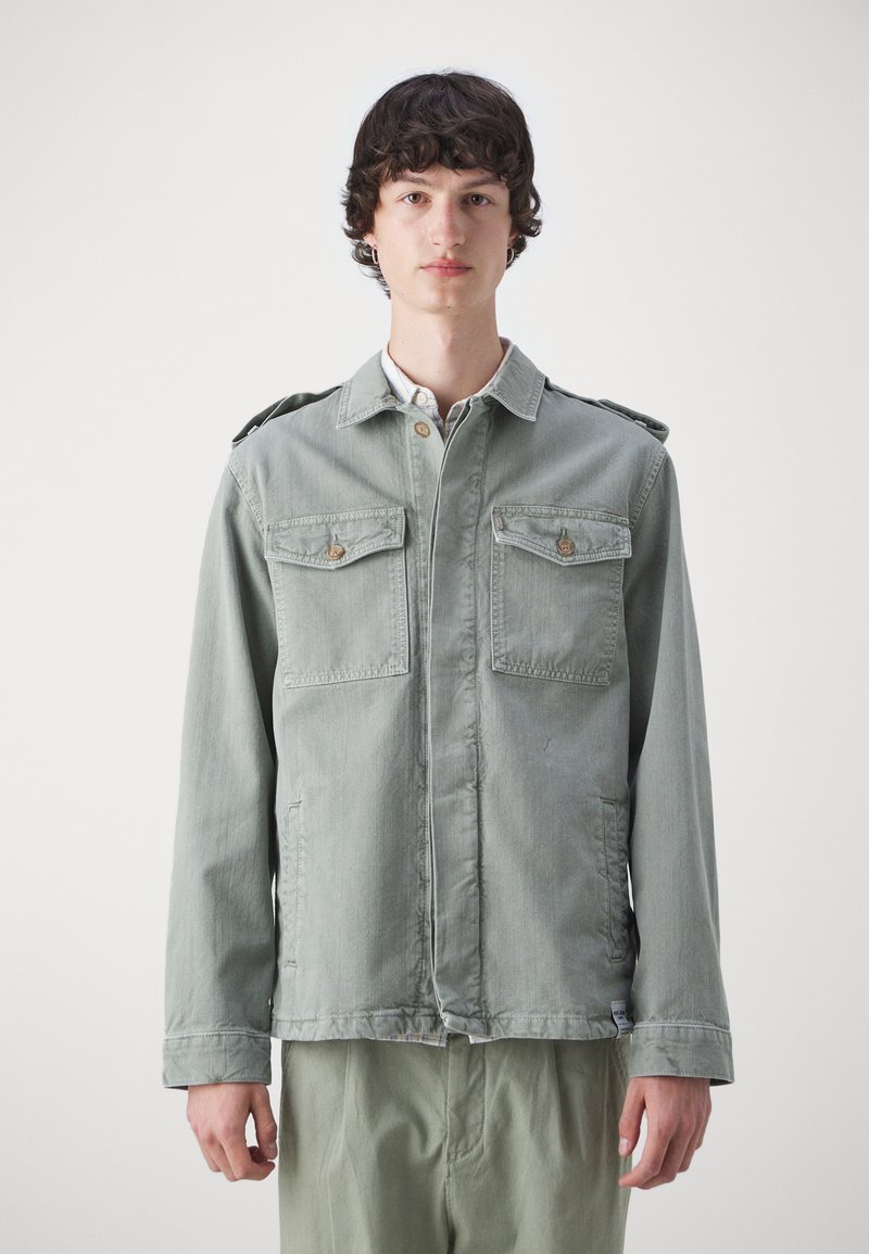 Chaqueta verde claro de estilo militar con dos bolsillos en el pecho, cierre de botones, hombreras y mangas largas. Textura de tela suave.