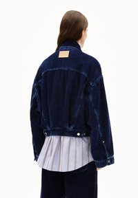 Giacca in corduroy blu navy con un design corto e oversized. Presenta un'etichetta rettangolare color sabbia sul retro e una chiusura a scatto in vita.