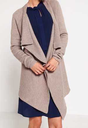 Frau trägt eine beige offene Strickjacke über einem marineblauen Kleid mit Schleifenkragen und steht vor einem schlichten weißen Hintergrund.