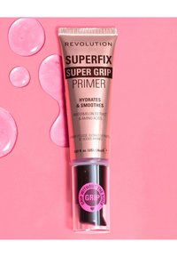 Revolution Superfix Super Grip Primer i en metallisk rosa tube med svarte aksenter, merket med fuktighetsgivende ingredienser. Glitrende rosa dråper i nærheten.