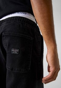 Joggers en denim noir avec ceinture élastique, ornés d'un patch logo Guess Jeans sur la poche arrière. La texture semble douce et décontractée.