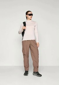 Lichtbeige langemouwen top met hoge kraag, gecombineerd met bruine cargobroek. Zwarte rugzak en zonnebril, met een gestructureerde stof.