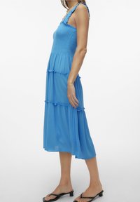 Robe midi à volants bleue en tissu léger. Dotée d'un corsage smocké, de bretelles à volants et d'une silhouette fluide. Portée avec des tongs noires.