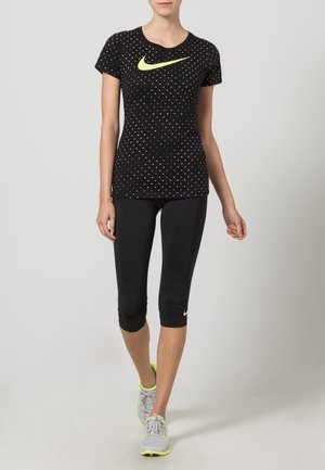 Camiseta deportiva negra con estampado, mangas cortas y un gran logo de Nike en neón amarillo, combinada con leggings capri negros y zapatillas grises.