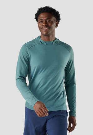 Lachende man loopt, draagt een teal lange mouwen hoodie en marineblauwe broek, tegen een effen lichte achtergrond.