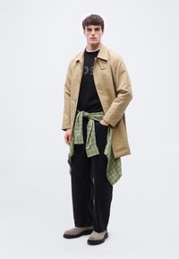 Νεαρός άνδρας που φοράει μπεζ παλτό πάνω από μαύρο πουκάμισο, πράσινο καρό πουκάμισο δεμένο στη μέση, μαύρο παντελόνι και γκρι μπότες, στέκεται μπροστά σε λευκό φόντο.