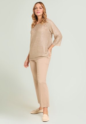 Vrouw staand met hand op de heup, gekleed in een beige gebreide trui, lichtbruine broek en crème loafers tegen een effen lichte achtergrond.