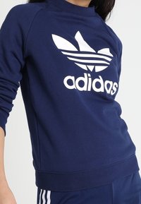 Sweatshirt bleu marine en matériau doux, avec un grand logo Adidas trèfle blanc et des poignets et un ourlet côtelés.