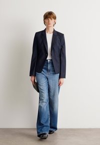 Námořnický pruhovaný blazer přes bílou košili, doplněný o světlé modré rozšířené džíny. Model drží černou kabelku. Minimalistický styl, jednoduché pozadí.