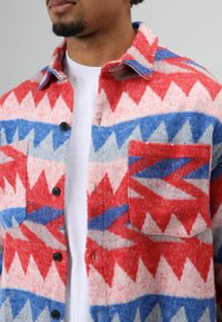 Rot, weiß und blau gemustertes Flanellhemd mit einem geometrischen Design, zwei Brusttaschen und schwarzen Knöpfen. Weiche Wolltextur.