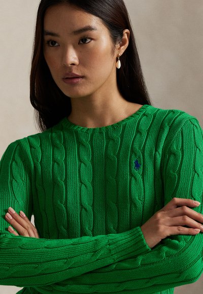 Tânără cu păr lung și închis la culoare purtând un pulover tricotat verde aprins, cu brațele încrucișate pe un fundal neutru.