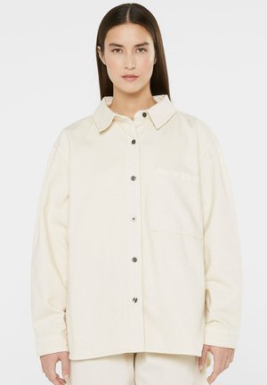 Woman with straight dark hair wearing oversized cream button-up shirt with front pocket and matching pants, facing forward against white background.