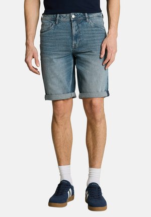 Mann trägt hochgekrempelte blaue Jeansshorts, weiße Kniestrümpfe und dunkelblaue Sneakers mit weißen Streifen, steht vor weißem Hintergrund.