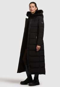 Schwarzer langer Puffer-Mantel mit einer mit Fleece gefütterten Kapuze, gestepptem Design, Frontreißverschluss und Seitentaschen. Getragen mit schwarzen Stiefeletten und einem beigen Kleid.