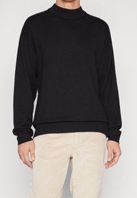 Zwarte gebreide trui met een hoge kraag, losse pasvorm, geribbelde mouwen en zoom. Gecombineerd met lichtbeige corduroy broek.