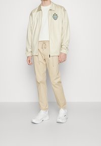 Béžový pruhovaný blazer s vyšívaným znakem, zkombinovaný s béžovými kalhotami na stažení a bílými teniskami. Hladká textura a volný střih.