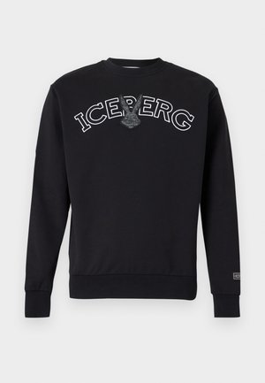 Mann trägt eine burgunderfarbene Kappe, schwarzen Sweatshirt mit dem Schriftzug „ICEBERG“, hellblaue Jeans mit Umschlägen und dunkelbraune Loafers, steht mit einer Hand in der Tasche.