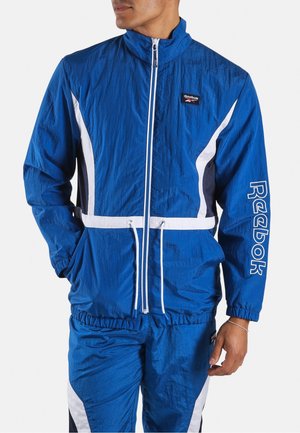 Mann trägt einen blauen Reebok-Trainingsanzug mit weißen und schwarzen Akzenten, Hände in den Taschen, steht vor einem einfarbigen Hintergrund.