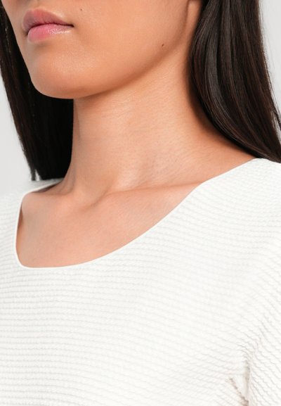Witte getextureerde top met een brede scoop halslijn en ribbeldetail, met gladde huid en donker stijlhaar zichtbaar bij de nek.